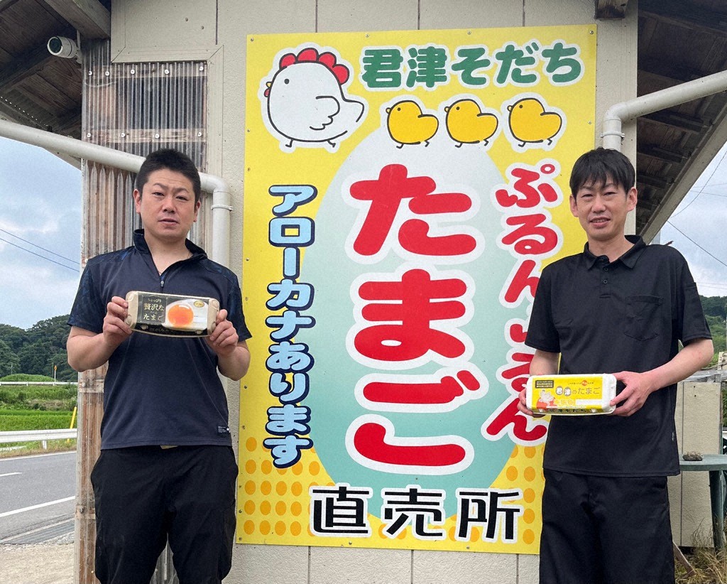 自動販売機によるたまご直売所の看板前に立つ靖友さん(右)と和喜さん