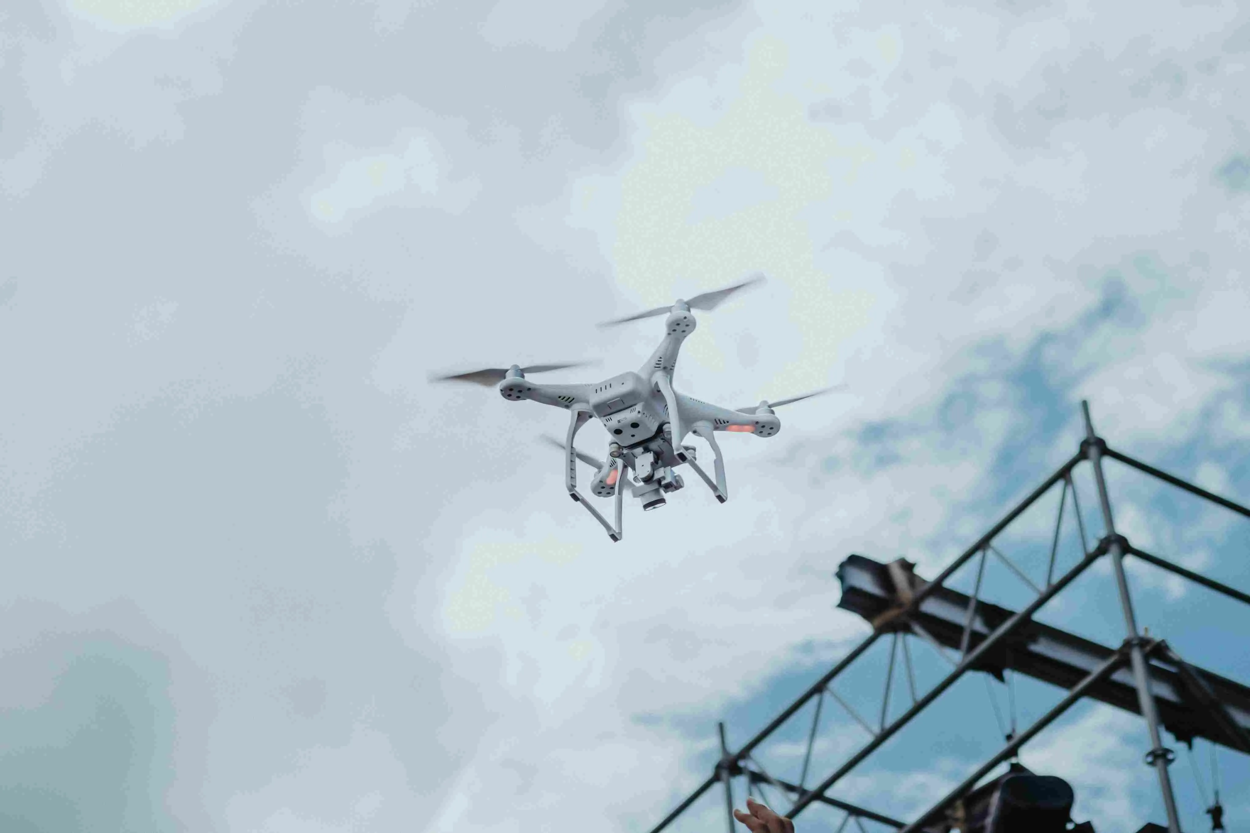A white quadcopter drone flying against a cloudy sky near a metal scaffolding structure, illustrating AI-powered autonomous technology and data collection in enterprise operations.