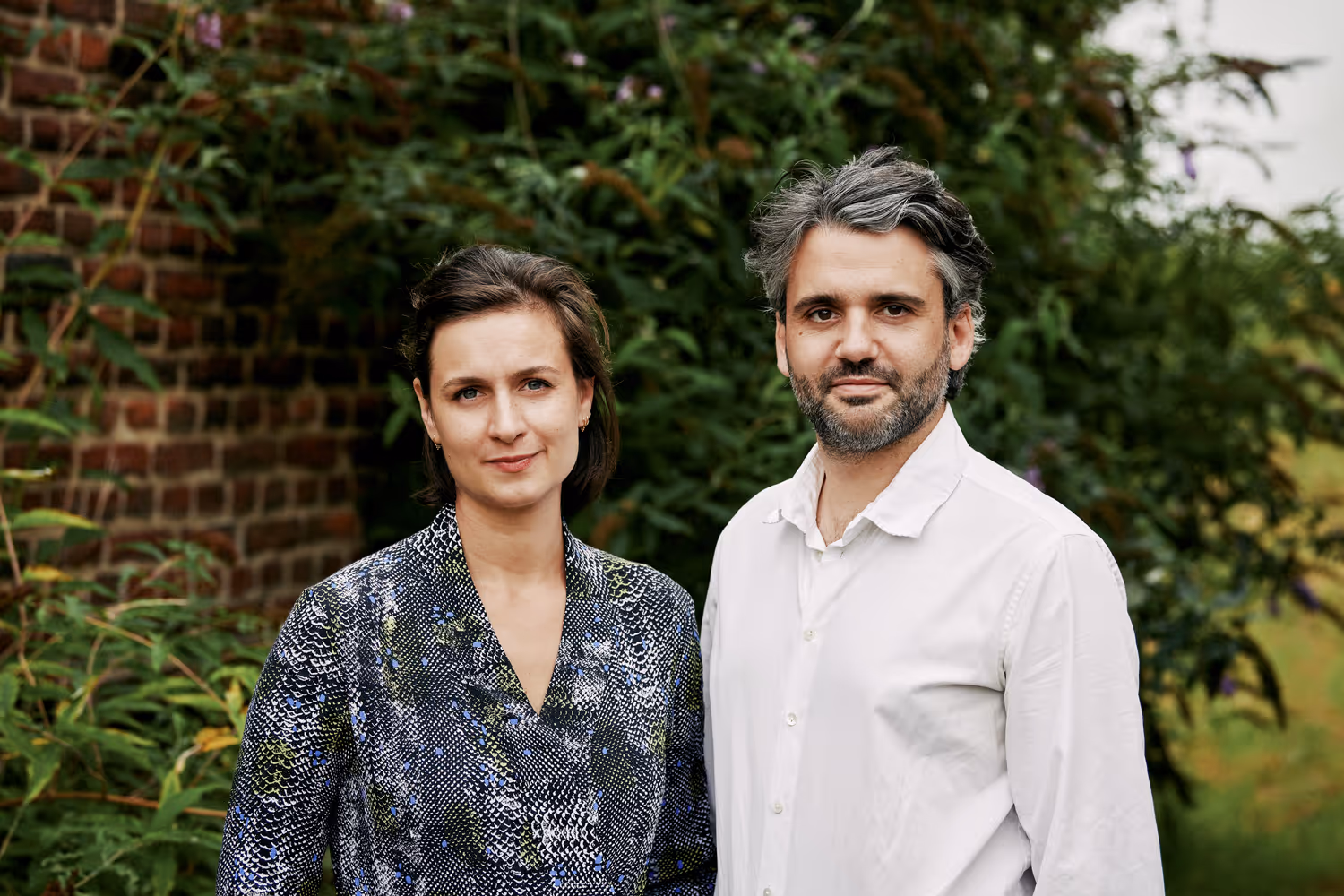Een man in een witte overhemd en een vrouw in een patroonblouse staan buiten met groene planten en een bakstenen muur op de achtergrond.