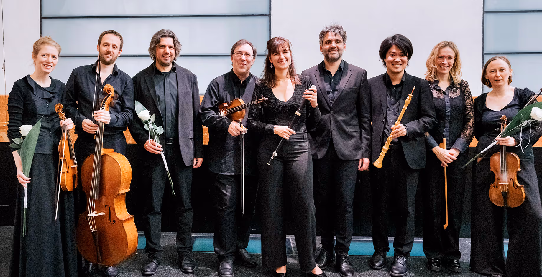 Groepsfoto van een muziekkwartet met acht mensen in formele kleding, sommigen met strijkinstrumenten en anderen met houten blaasinstrumenten en bloemen in hun handen.
