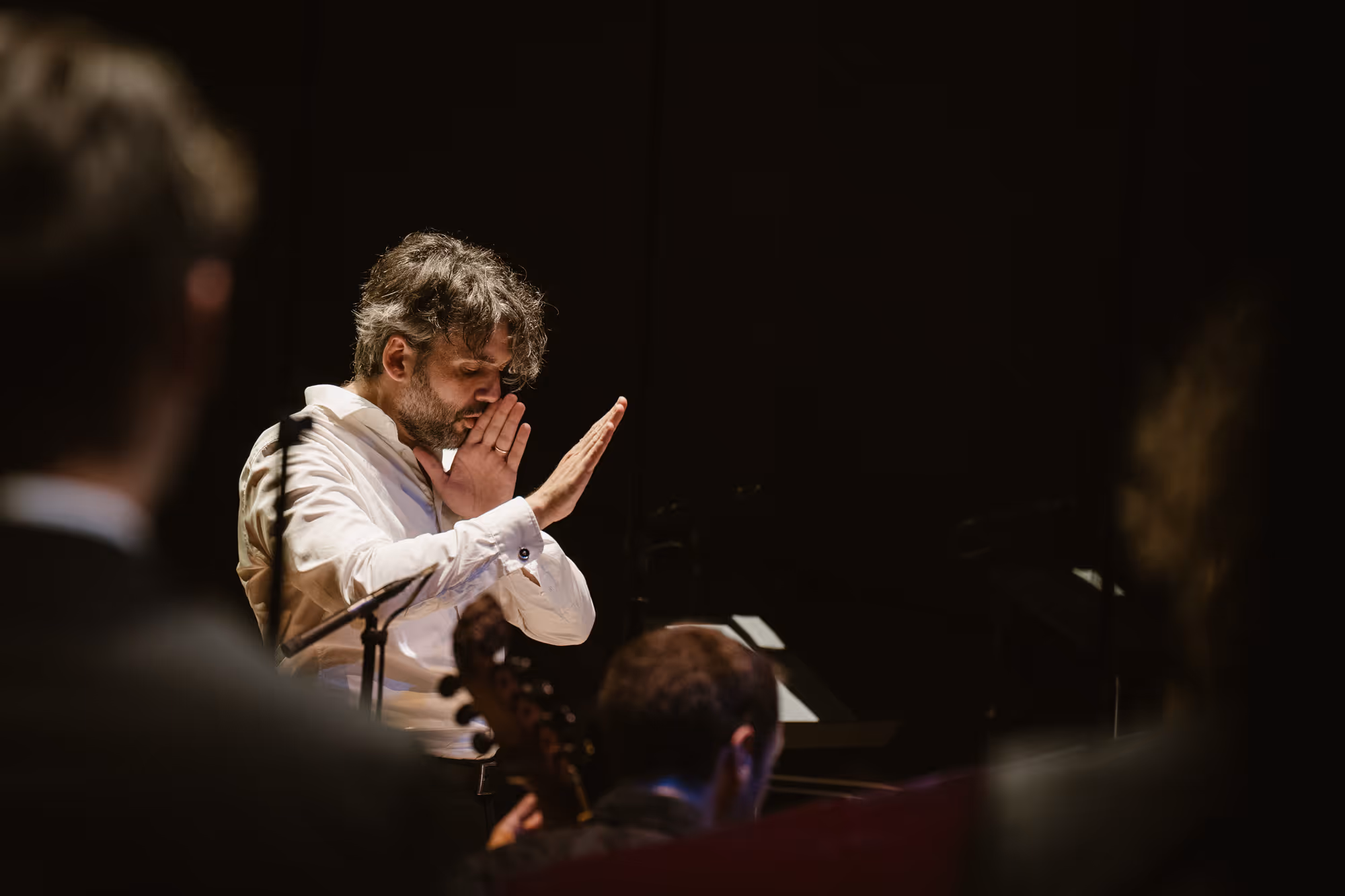 Man in witte blouse dirigeert een orkest in een donkere ruimte met geconcentreerde blik.