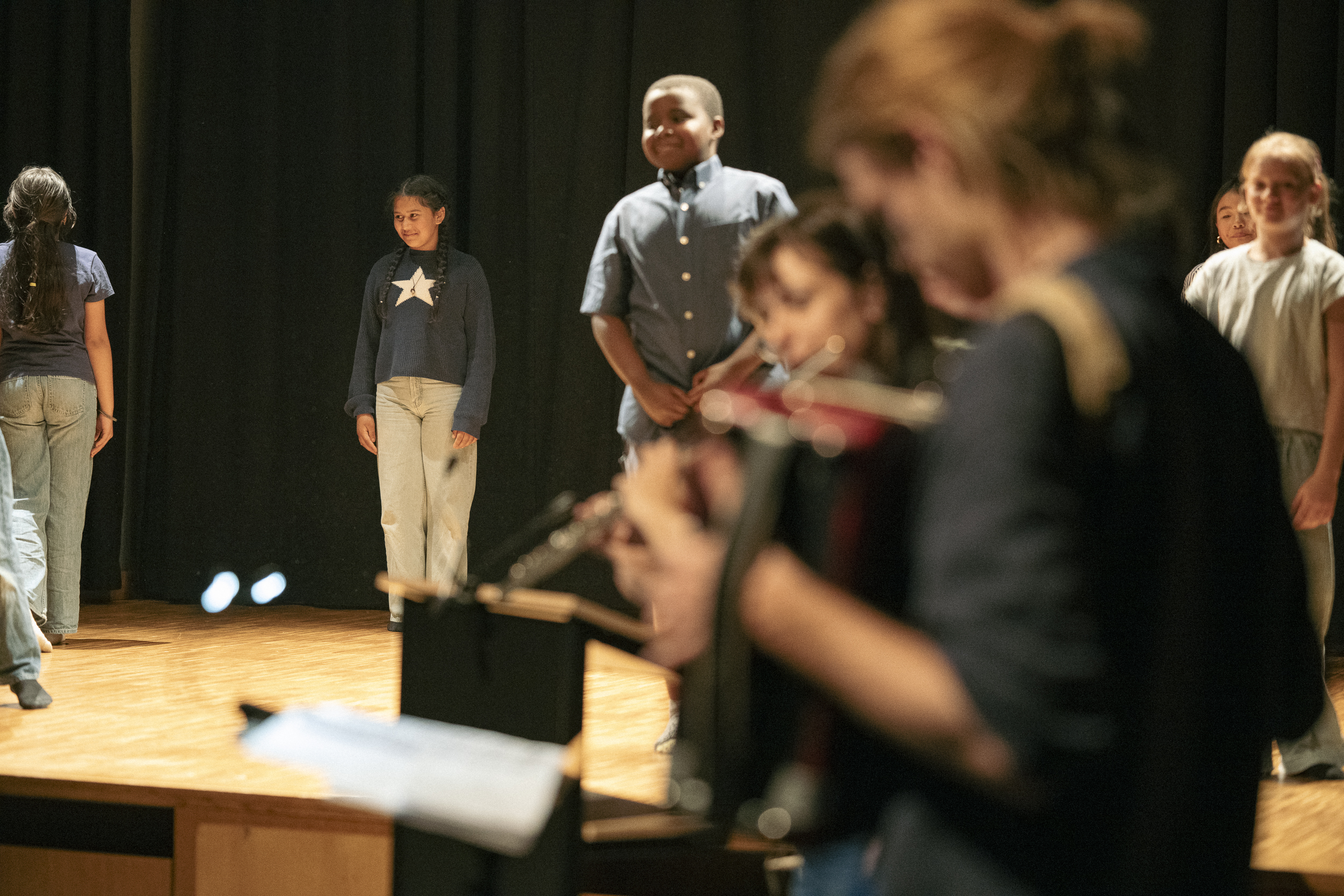 Orkest met muzikanten die strijkinstrumenten bespelen, met dirigent voor het podium en publiek dat toekijkt in een concertzaal.