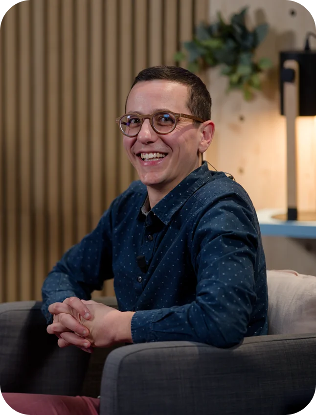 Portrait de Damien Fournet de profil, assis dans un fauteuil gris, tourné vers la gauche, portant une chemise bleue et des lunettes et souriant