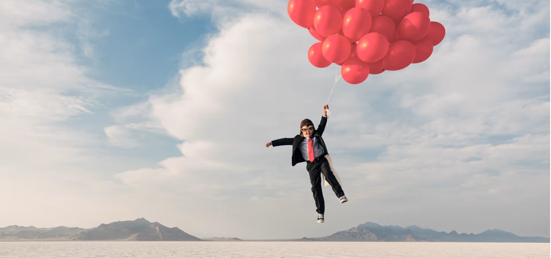 Ein Kind im Anzug mit roter Krawatte und Pilotenbrille fliegt, gehalten von vielen roten Luftballons, über eine weite, trockene Landschaft mit Bergen im Hintergrund.