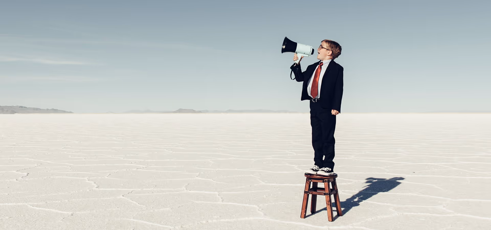 Ein Junge im Anzug steht auf einem Hocker in einer weiten weißen Landschaft und spricht in ein Megafon.