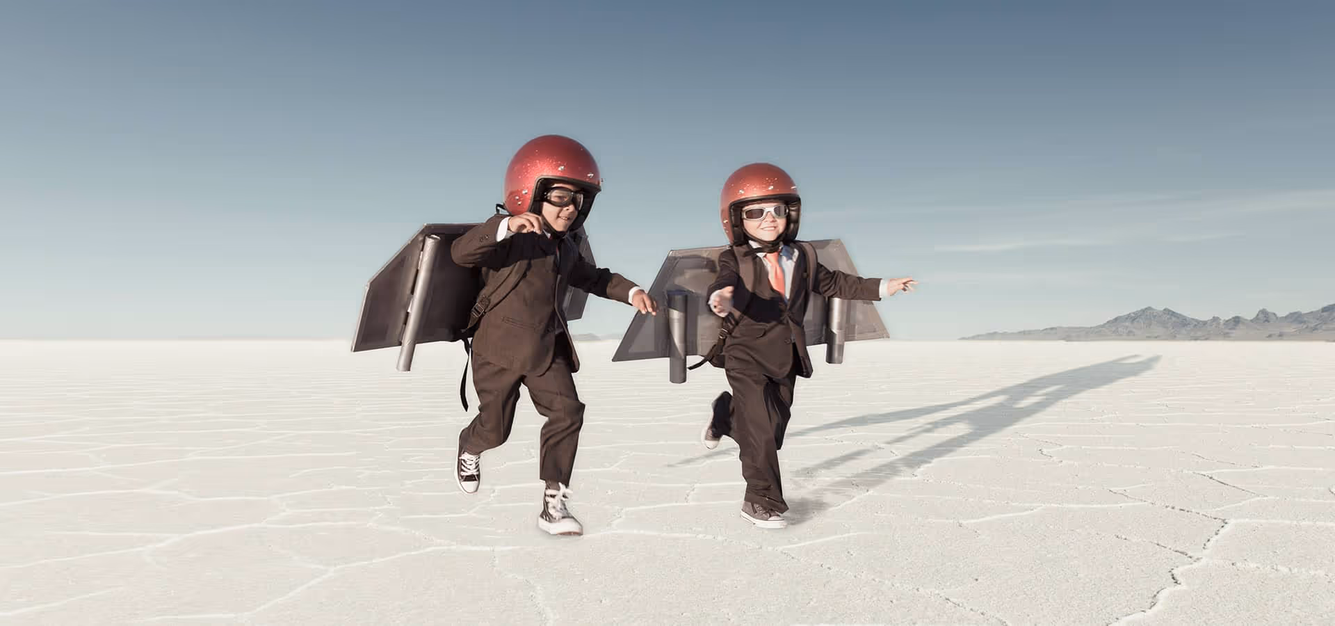 Zwei Kinder in Anzügen und roten Helmen rennen auf einer trockenen Salzebene mit selbstgebastelten Flügeln auf dem Rücken.