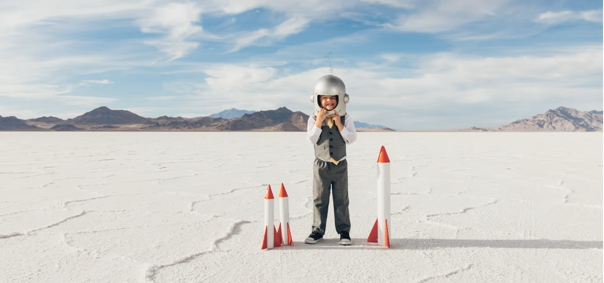 Ein Kind in James-Bond-Anzug und Weltraumhelm steht lächelnd auf einer weißen Salzebene zwischen drei kleinen Raketenmodellen.