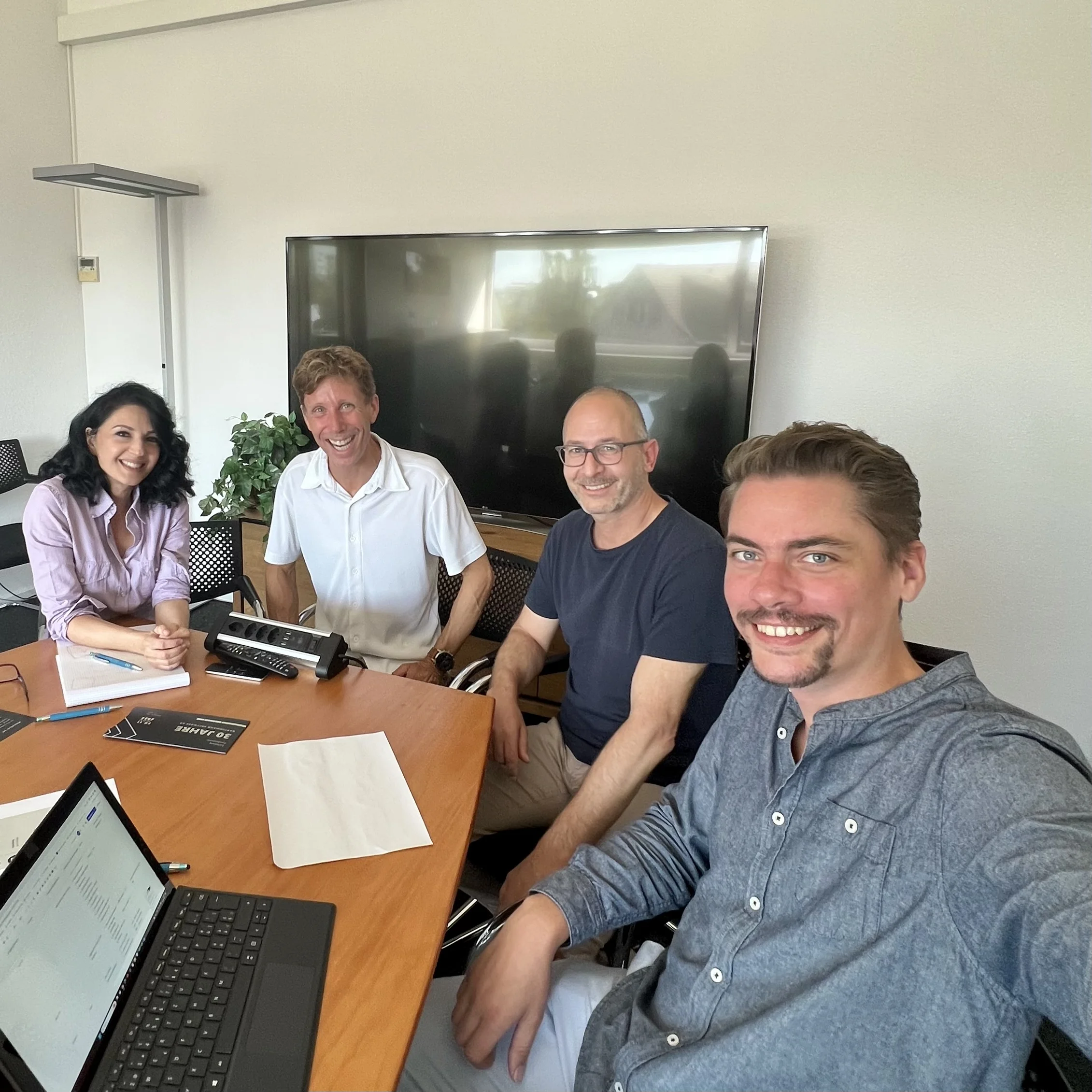Vier Personen sitzen um einen hölzernen Konferenztisch in einem Büro, mit einem Laptop, Notizblock und Bildschirm hinter ihnen.