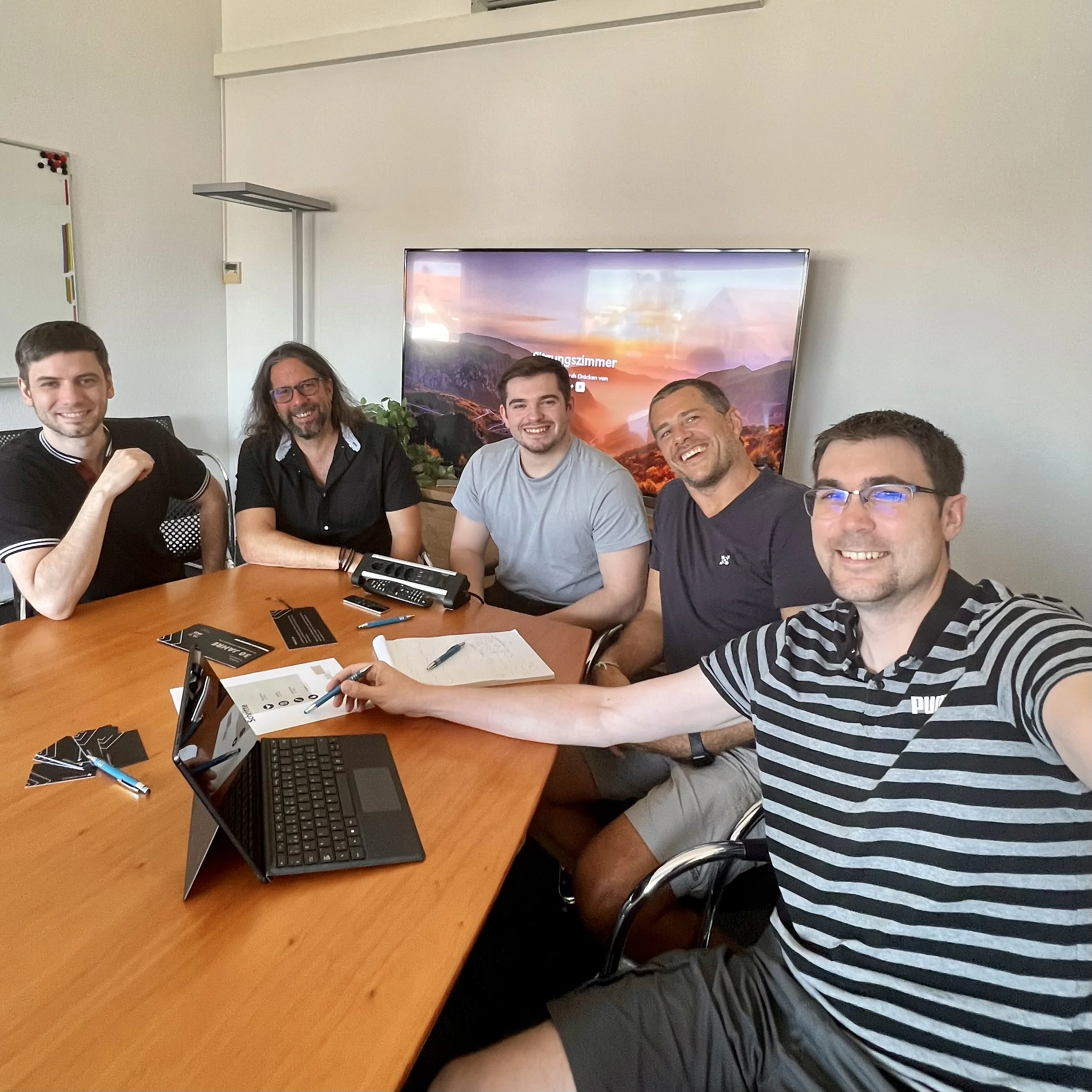 Fünf Männer sitzen um einen Konferenztisch mit Laptop, Notizen und Stiften und lächeln in einem Büro.