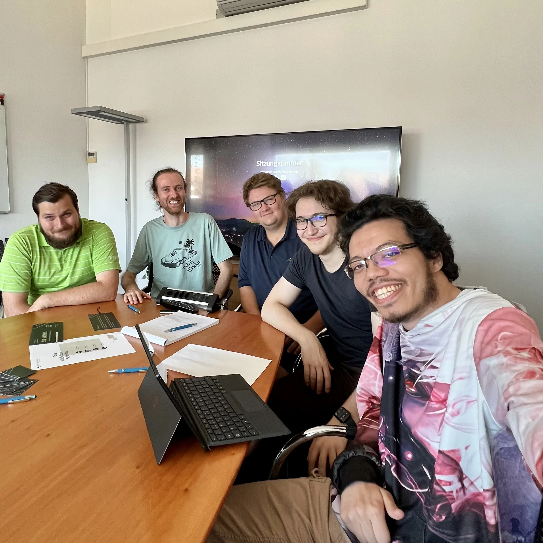 Fünf Personen lächeln bei einem Treffen an einem Konferenztisch mit Laptops und Papieren.