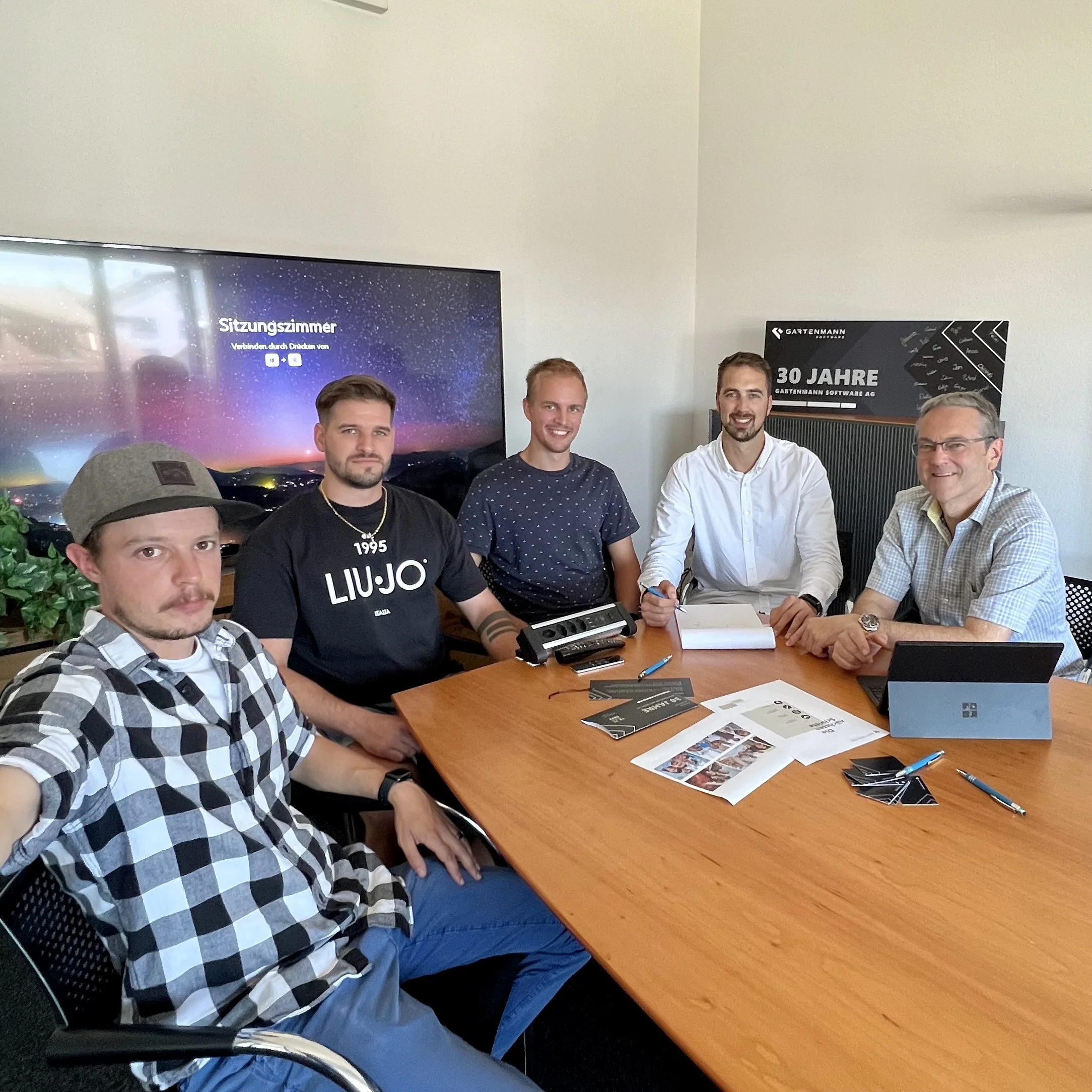 Fünf Männer sitzen an einem Besprechungstisch mit Unterlagen, Tablet und Stiften in einem Büro mit großem Bildschirm im Hintergrund.