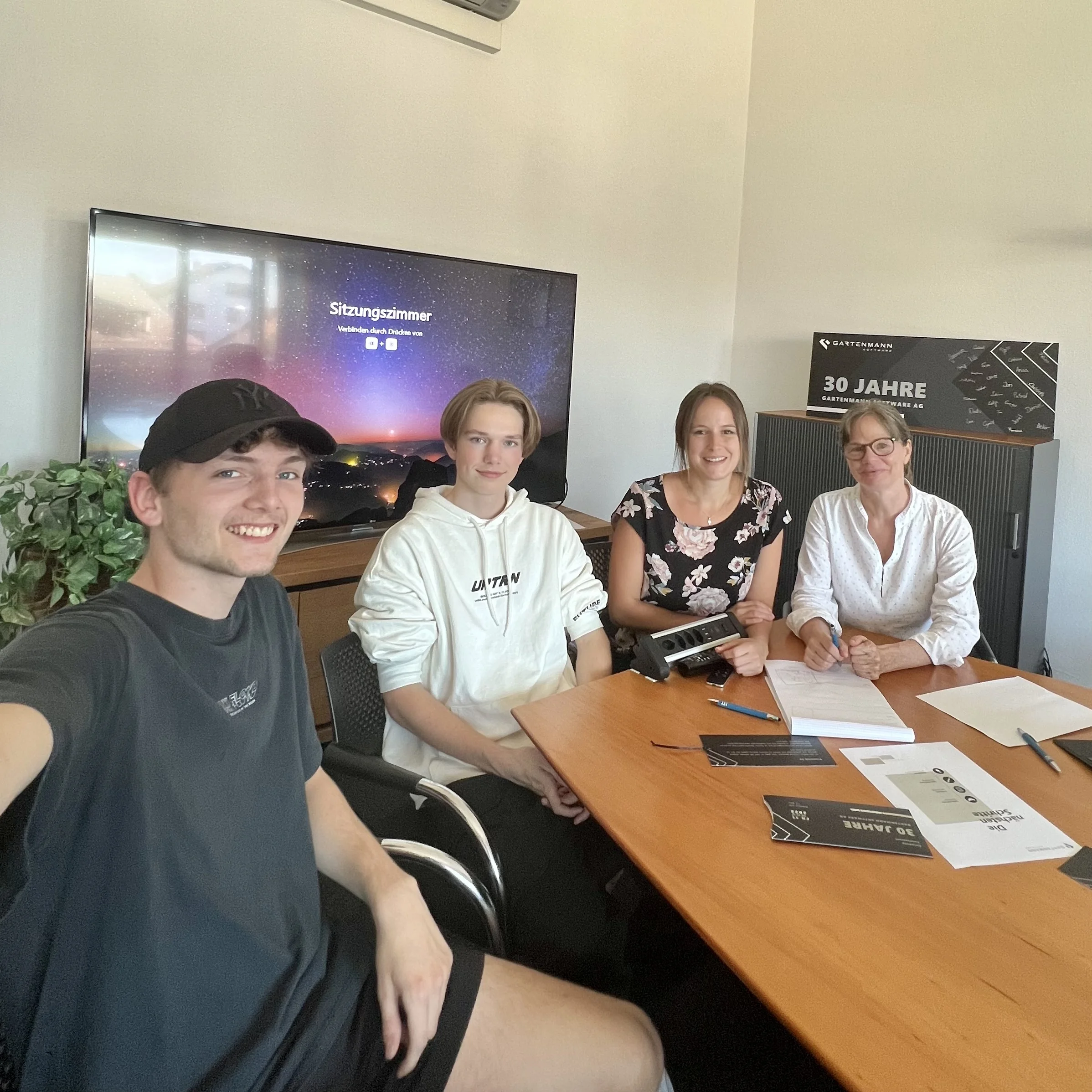 Vier Personen sitzen an einem Besprechungstisch in einem hellen Büro mit Dokumenten und Stiften auf dem Tisch.