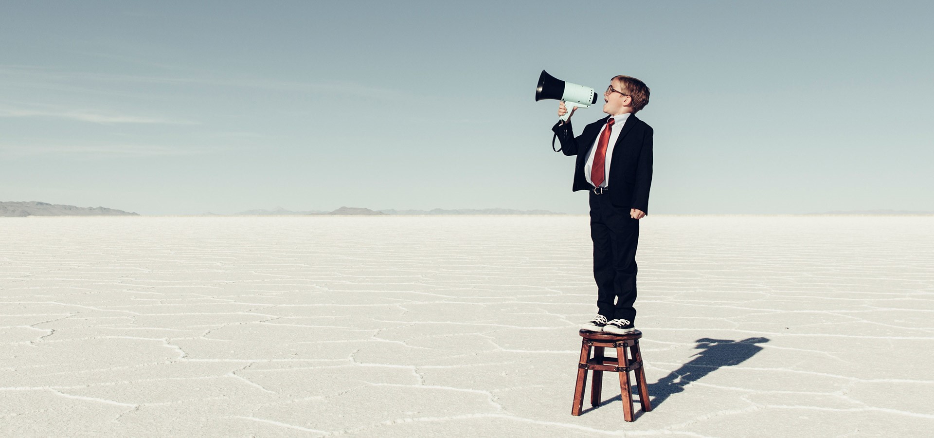 Junge im Anzug steht auf einem Hocker in einer weiten, salzverkrusteten Landschaft und spricht in ein Megafon.