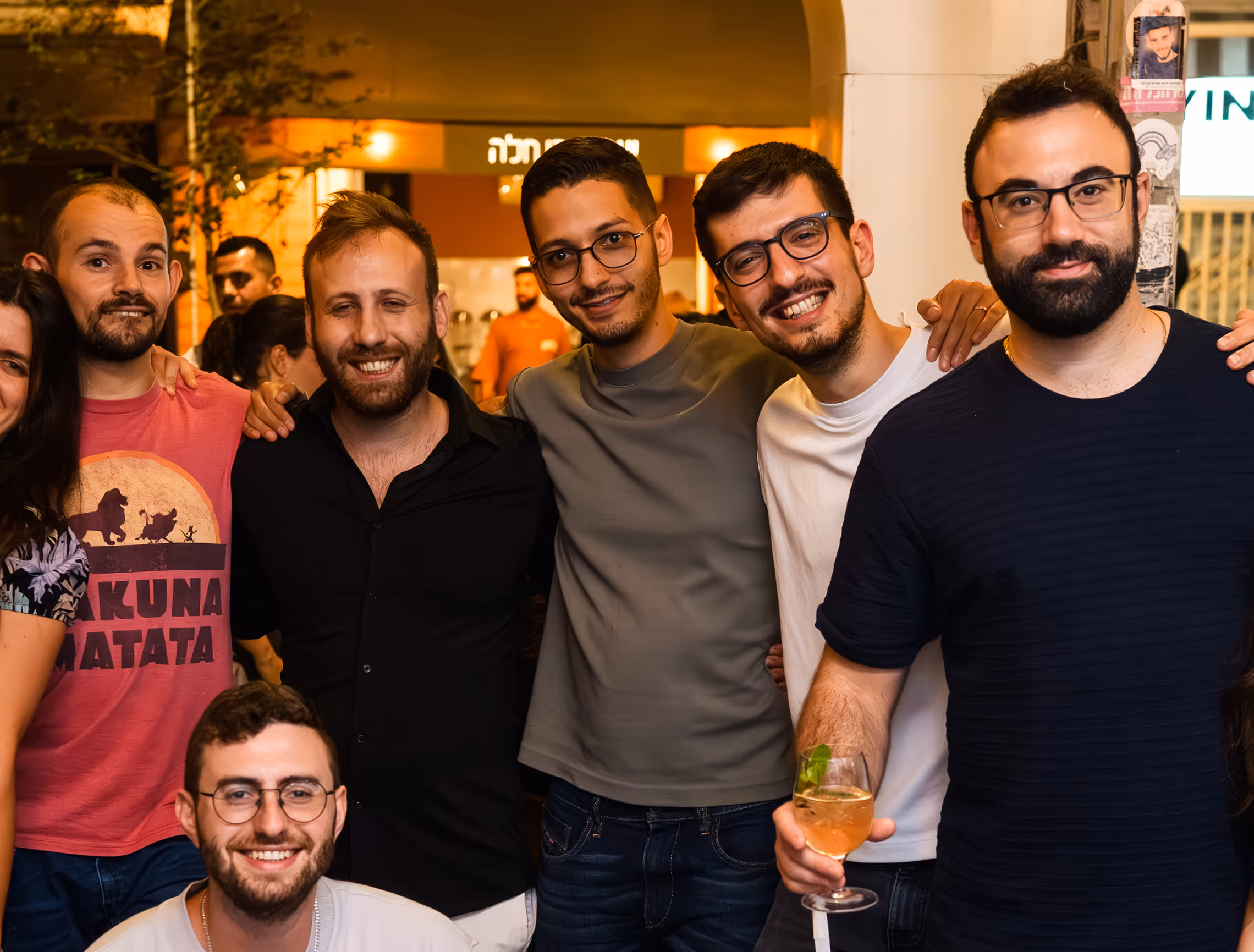 Group of six men smiling and posing closely together indoors, one holding a drink.