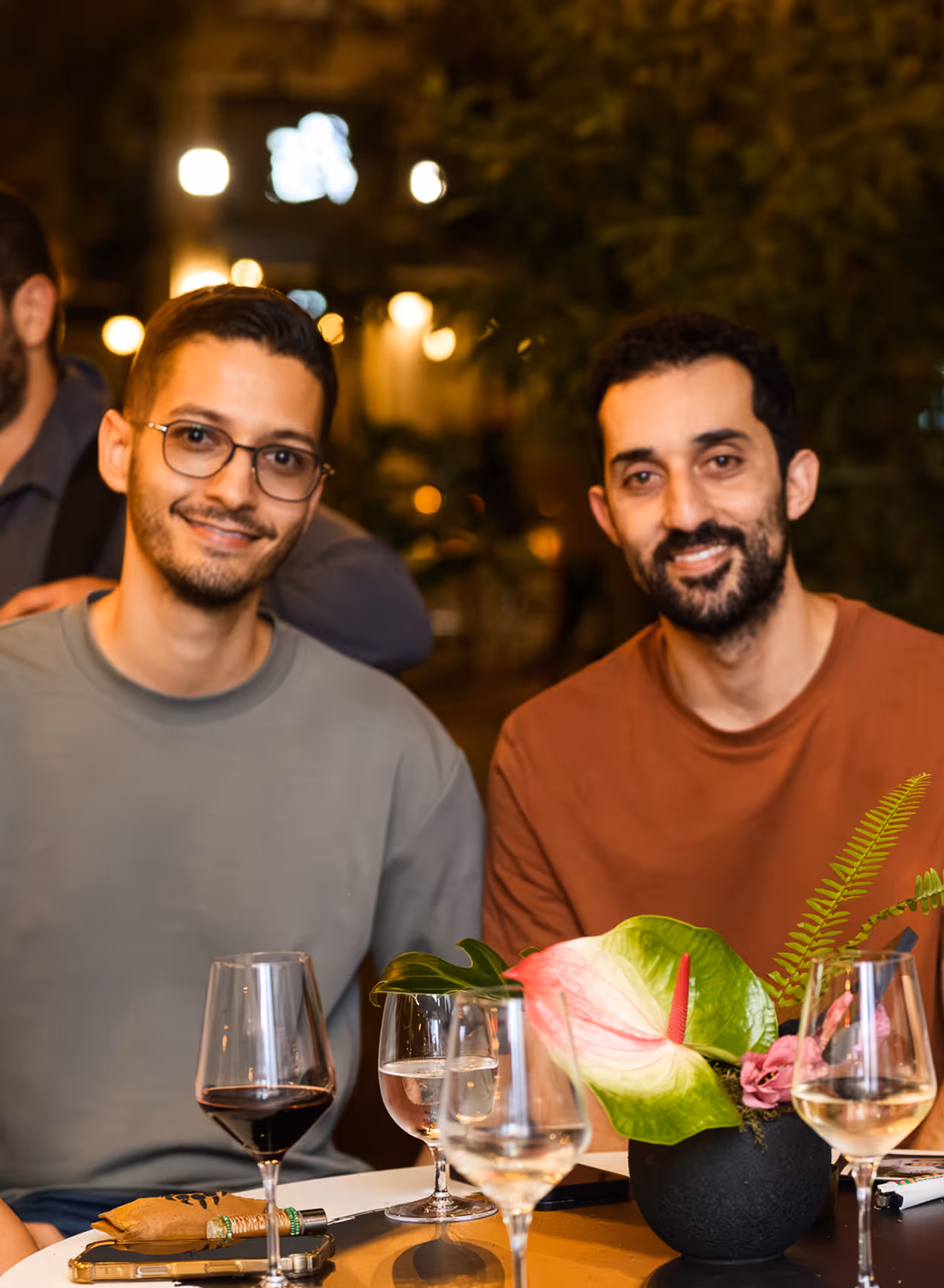 Two men smiling and sitting at a table with wine glasses and a vase with green and pink flowers in a warmly lit setting.