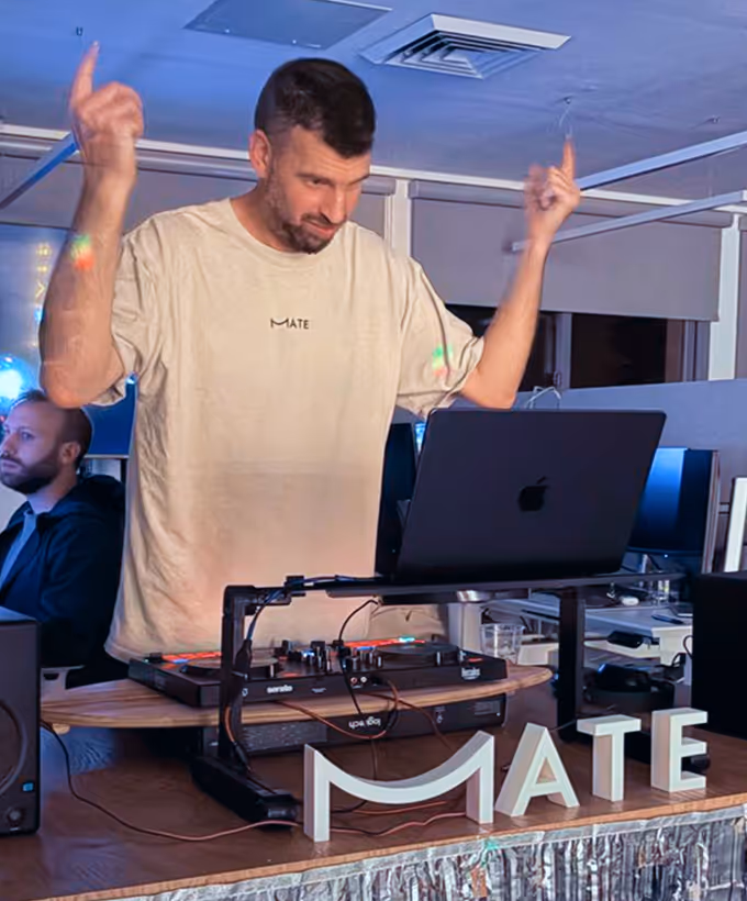Man DJing indoors with laptop and mixer on a wooden table displaying the word MATE.