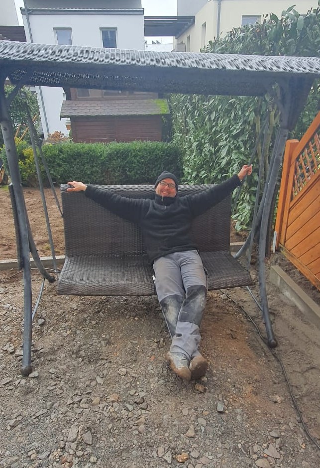 Person in black jacket and dirty pants sitting with arms outstretched on an outdoor swing bench.