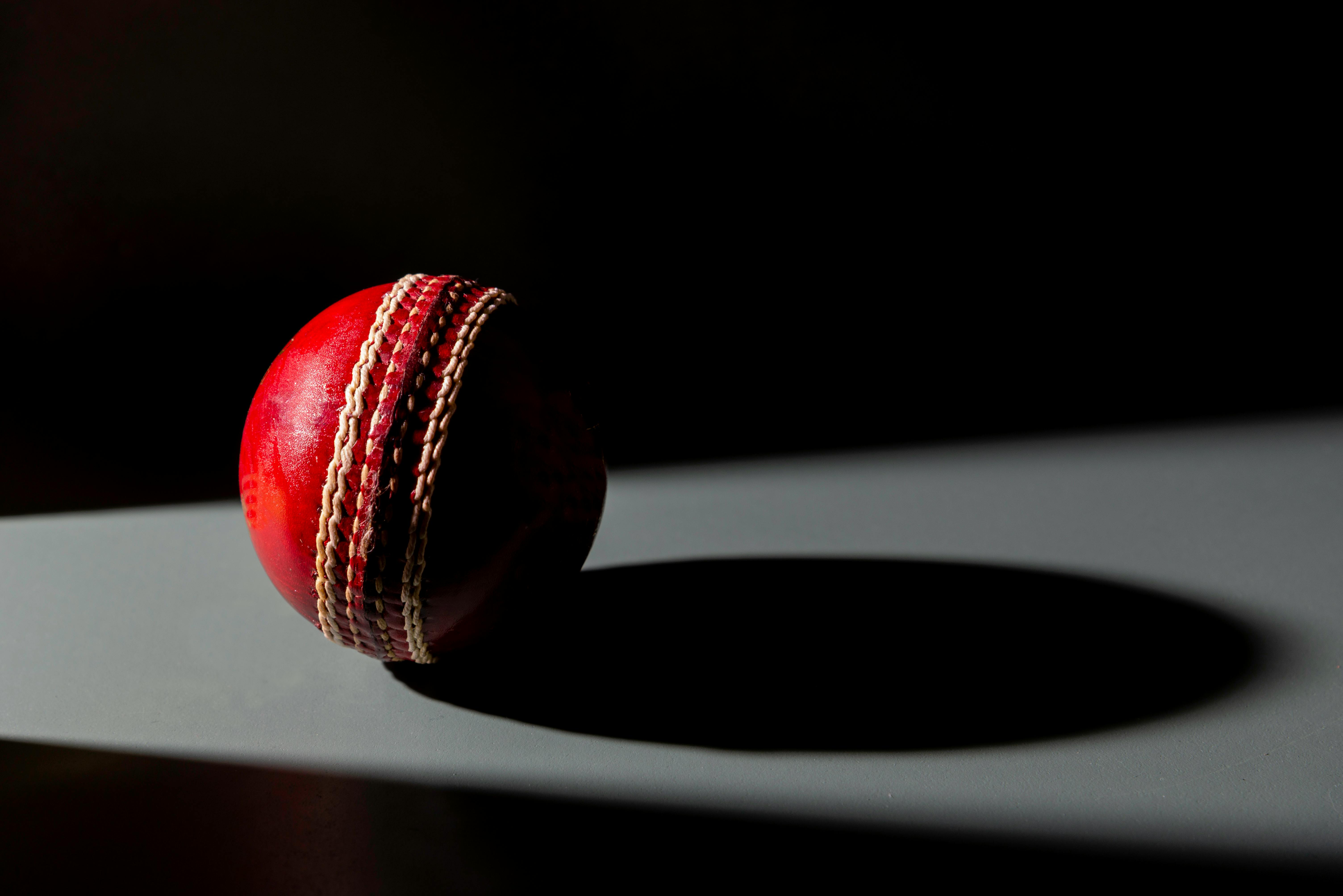Red cricket ball casting a shadow on a gray surface with a dark background.