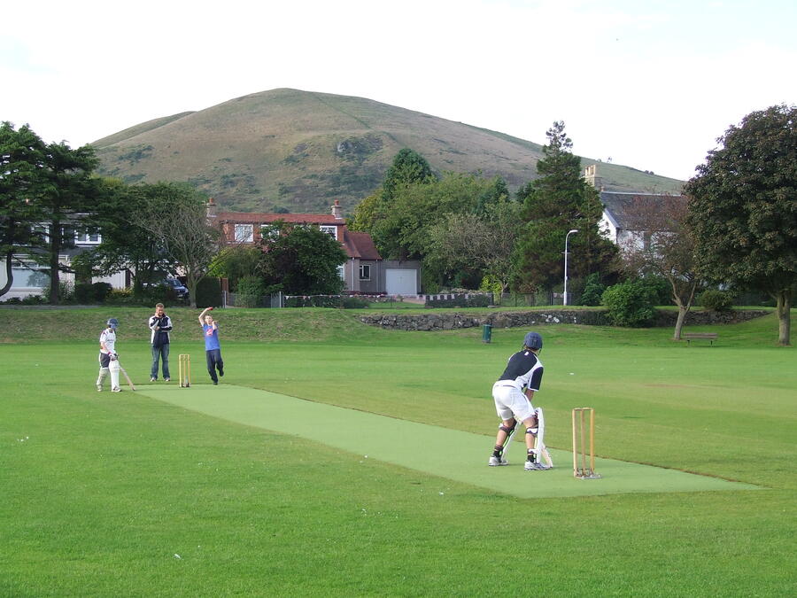 Four children playing cricket on a green field with a hill and houses in the background.