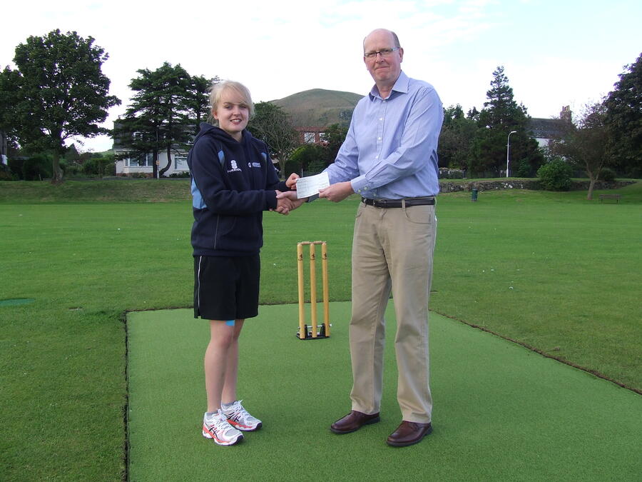 A young woman in sportswear and a man in business casual clothes shaking hands and exchanging a check on a cricket pitch.
