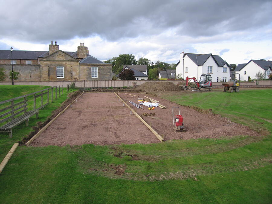 Construction site with leveled soil and wooden framing in a grassy area near residential houses and an old stone building under cloudy sky.