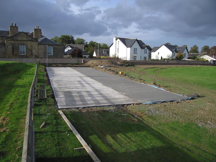 Long green lawn with a newly poured concrete slab in the center surrounded by residential houses under a cloudy sky.