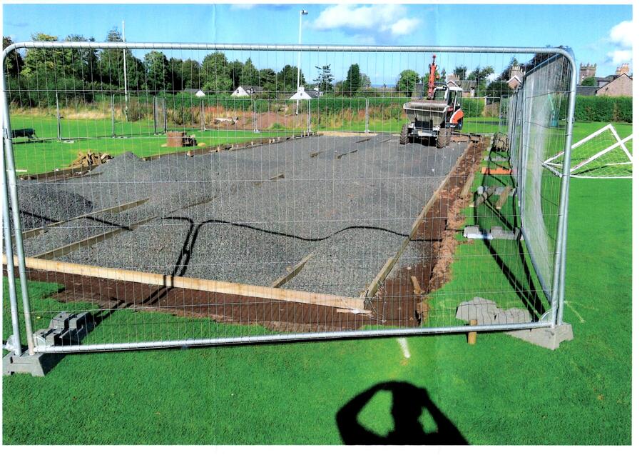 Construction site on green field enclosed by metal fencing with gravel spread over leveled ground and a small vehicle in the background.