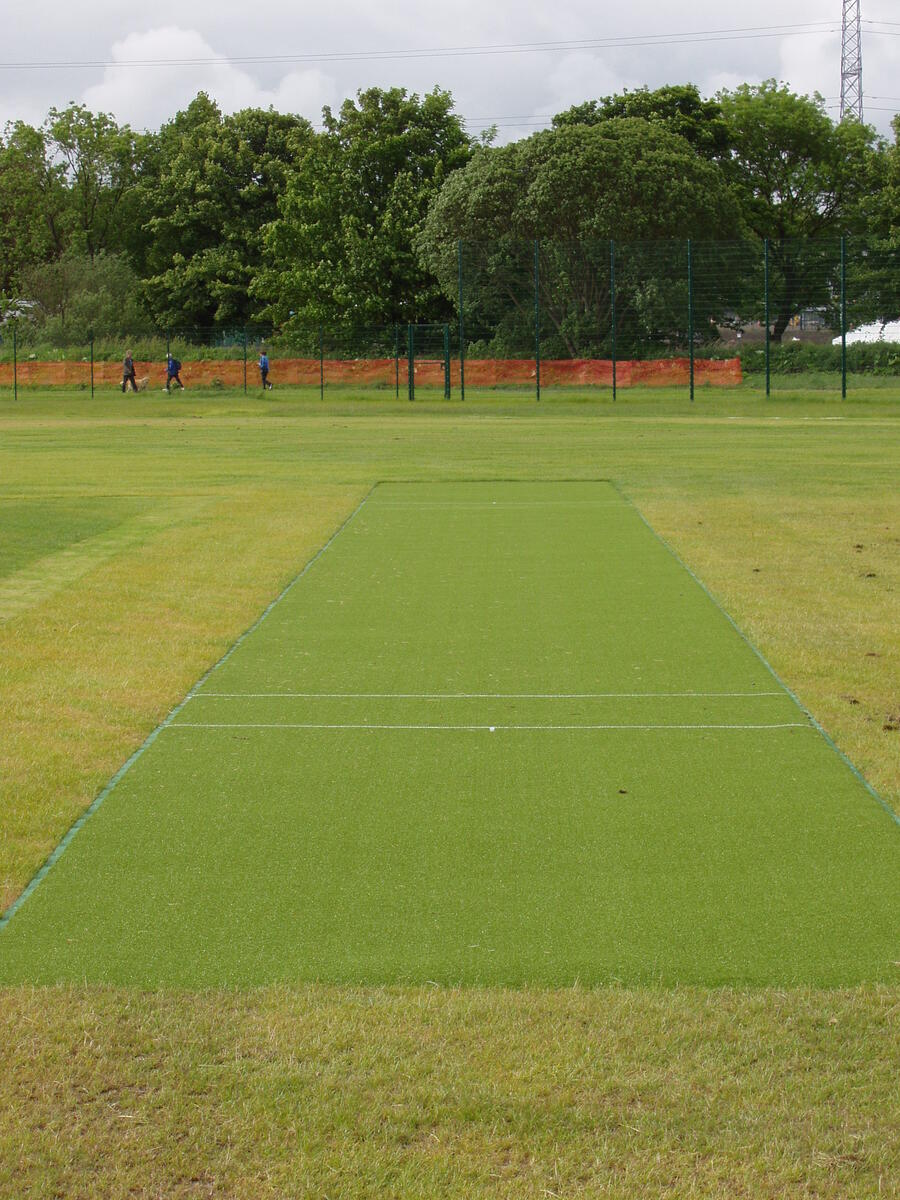 Artificial cricket pitch center on a grassy field with trees and three people walking in the background.