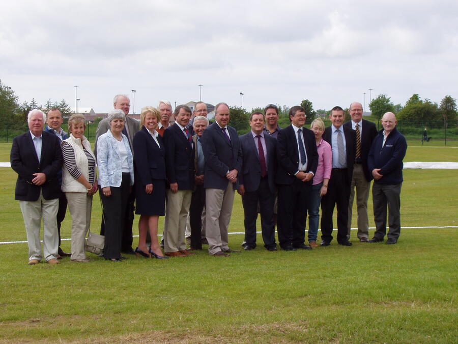 Group of sixteen adults standing outdoors on a grassy field, dressed mostly in formal and business casual attire.