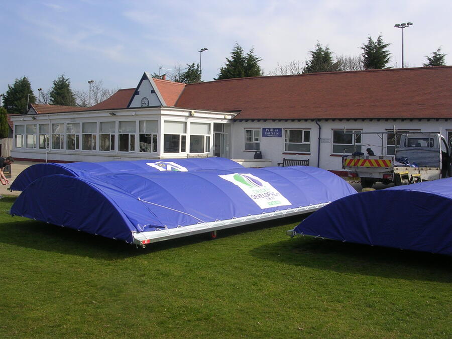 Blue portable cricket covers on green grass in front of a white pavilion building with red roof.