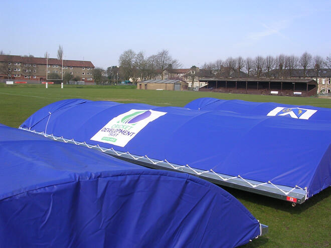 Blue covered portable cricket pitch covers set on grass near residential buildings and a cricket pavilion.