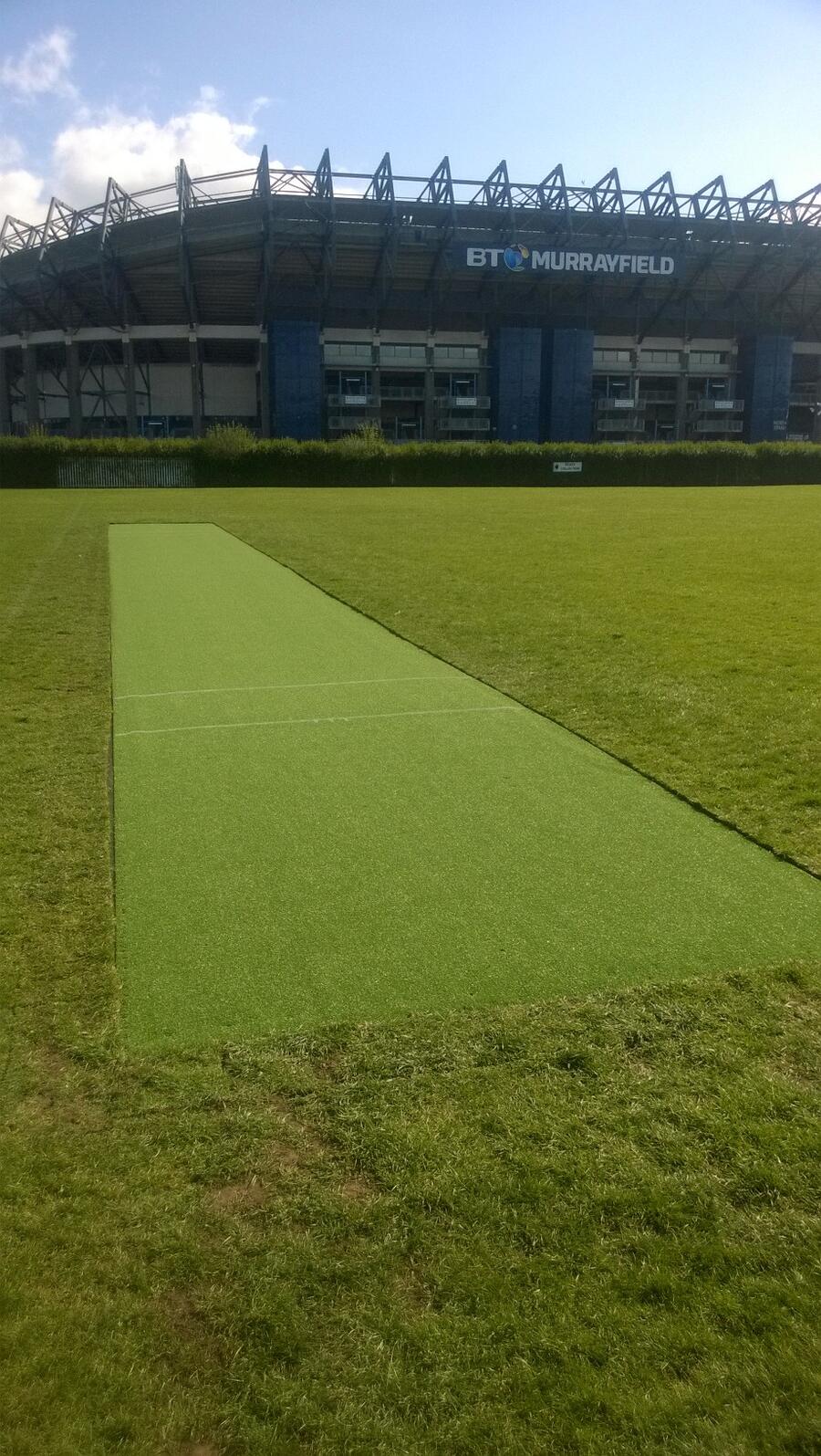 Cricket pitch with lush green grass in front of the BT Murrayfield stadium under a blue sky.