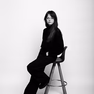 Jeune femme aux longs cheveux assise sur un tabouret, vêtue d'un pull noir et portant des lunettes.