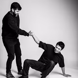 Un homme debout aide un autre homme à se relever en lui tendant la main, tous deux souriants en noir et blanc.