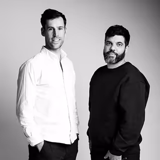 Portrait en noir et blanc de deux hommes debout, l'un en chemise blanche et l'autre en pull noir.