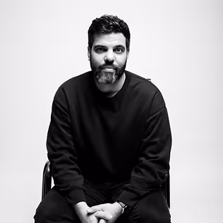 Portrait en noir et blanc d'un homme à barbe, assis, portant un pull foncé, regardant vers l'avant avec une expression neutre.