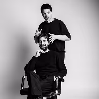 Un homme barbe assis souriant avec un autre homme debout derrière lui faisant un geste ludique avec ses mains.