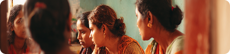 Four women engaged in a focused discussion, sitting closely together indoors with one woman partially visible in a mirror.