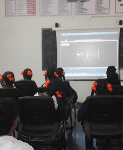 Students with orange hair bows sitting in chairs facing a classroom projector screen displaying a graph.