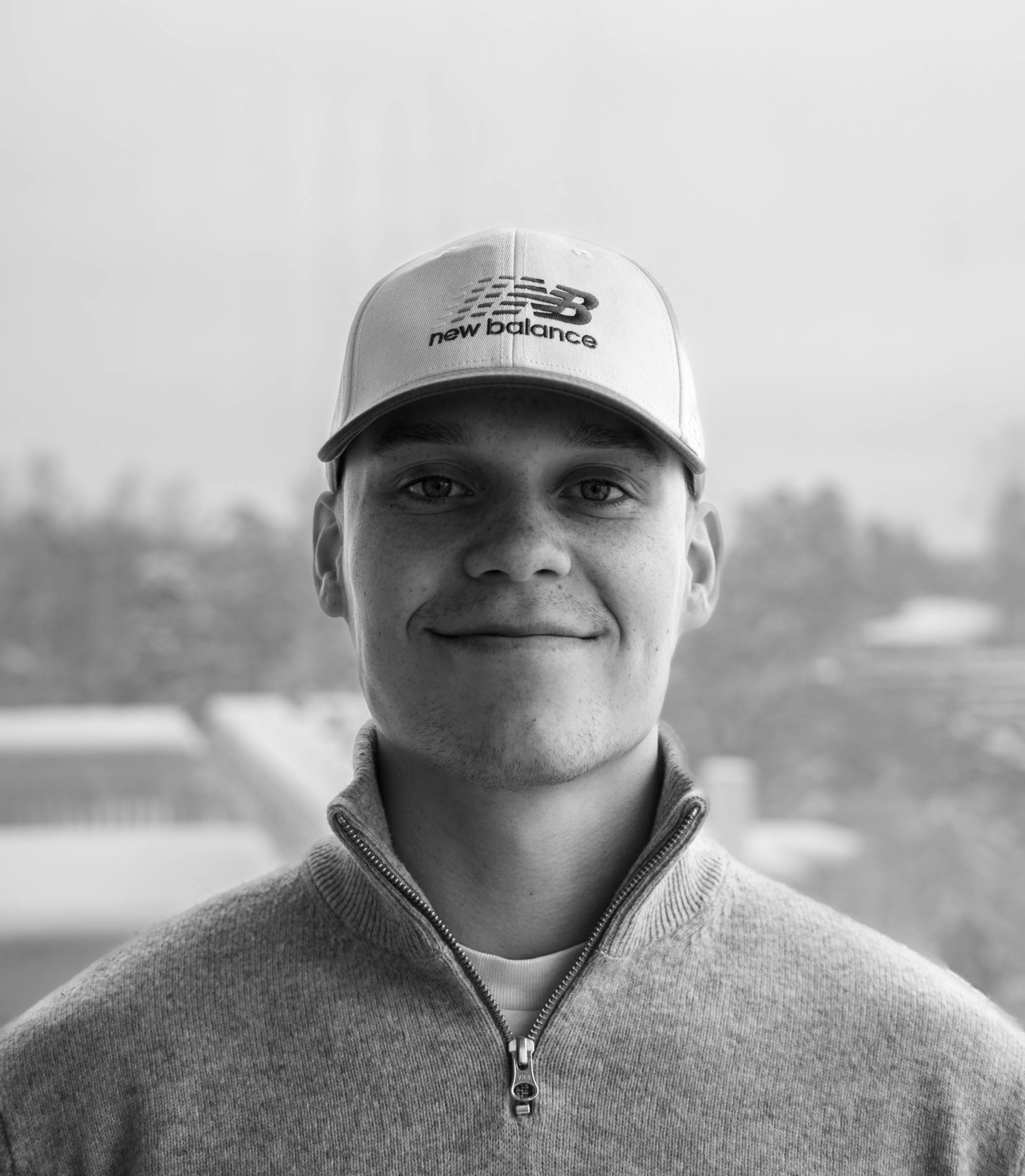 Smiling young man wearing a New Balance baseball cap and a zip-up sweater, with a blurred outdoor background.