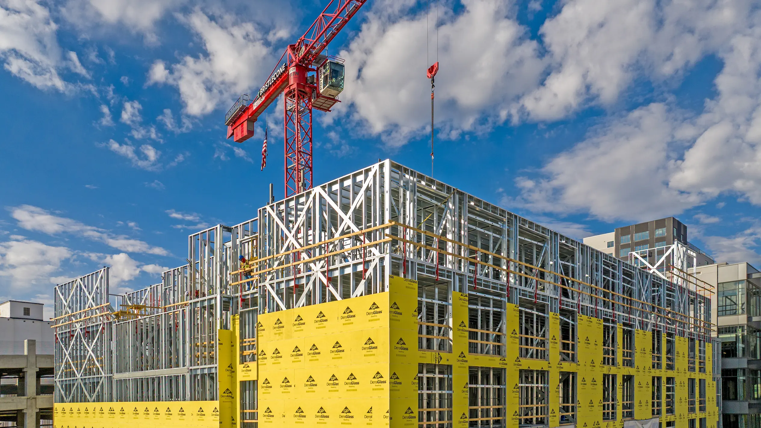 Under-construction multi-story building with steel framework and yellow sheathing panels, a red crane overhead, and a partly cloudy blue sky.
