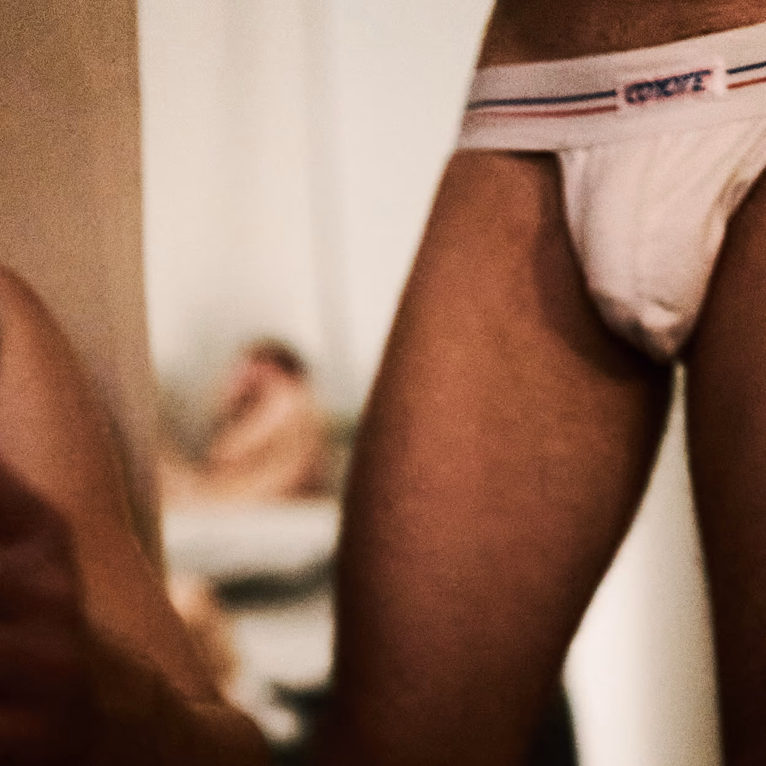 Vue rapprochée sur les jambes d'un homme en slip blanc avec bande tricolore, avec une autre personne floue allongée en arrière-plan.