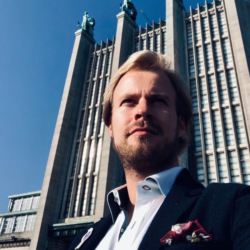 Corner storefront of a clothing shop with a beige coat on a mannequin displayed in the window and colorful garments hanging inside.