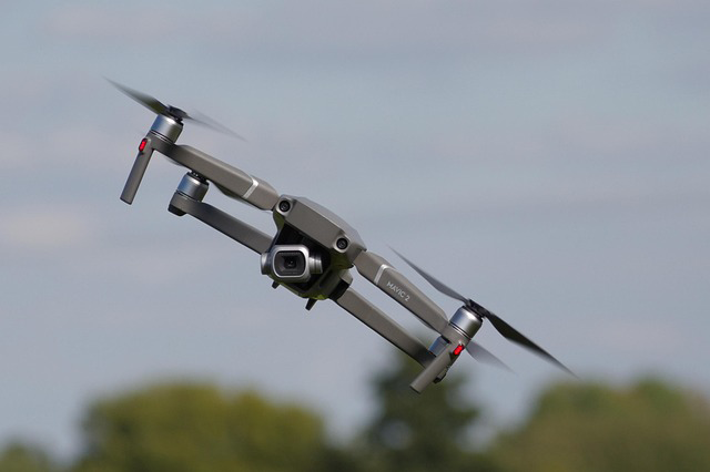 Drone gris à quatre hélices volant avec un arrière-plan flou d'arbres et ciel.