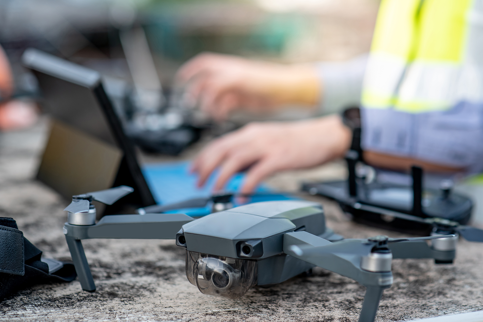 Un drone gris posé sur une surface avec un opérateur à l'arrière-plan utilisant un ordinateur portable.