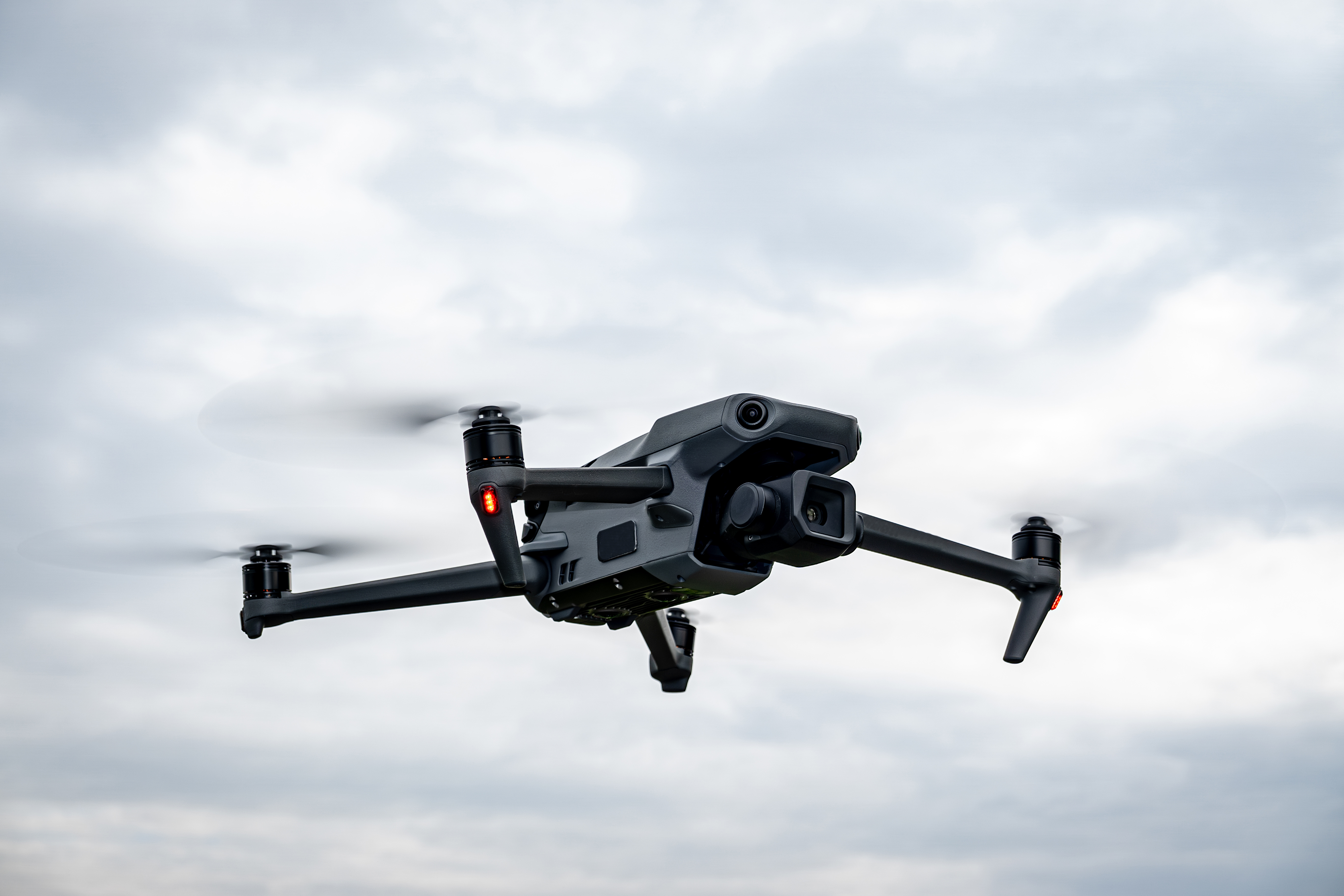 Drone noir en vol avec des hélices en rotation et un ciel nuageux en arrière-plan.