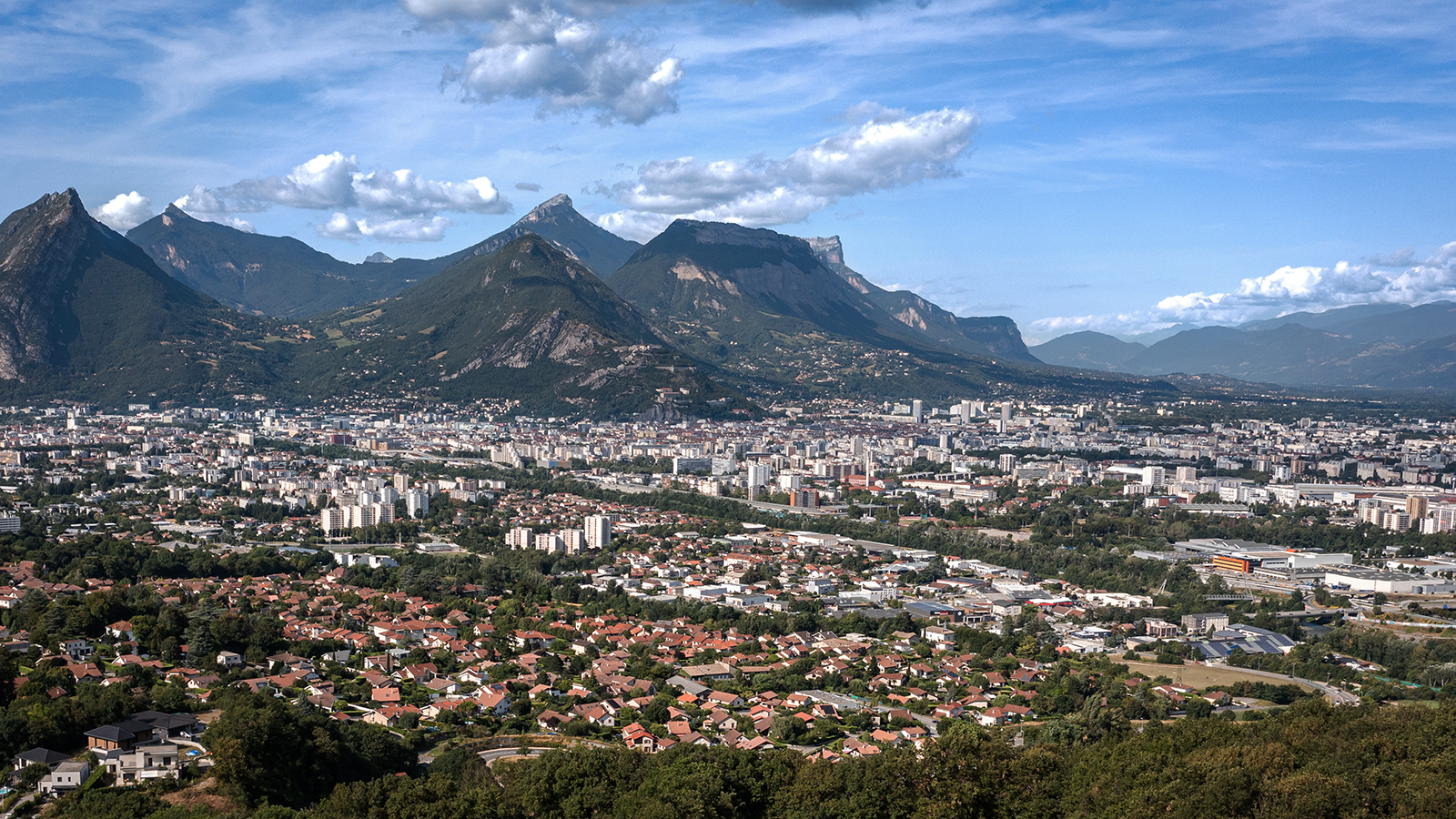 Vue panoramique d’une ville avec des maisons, des immeubles, et des montagnes verdoyantes en arrière-plan.