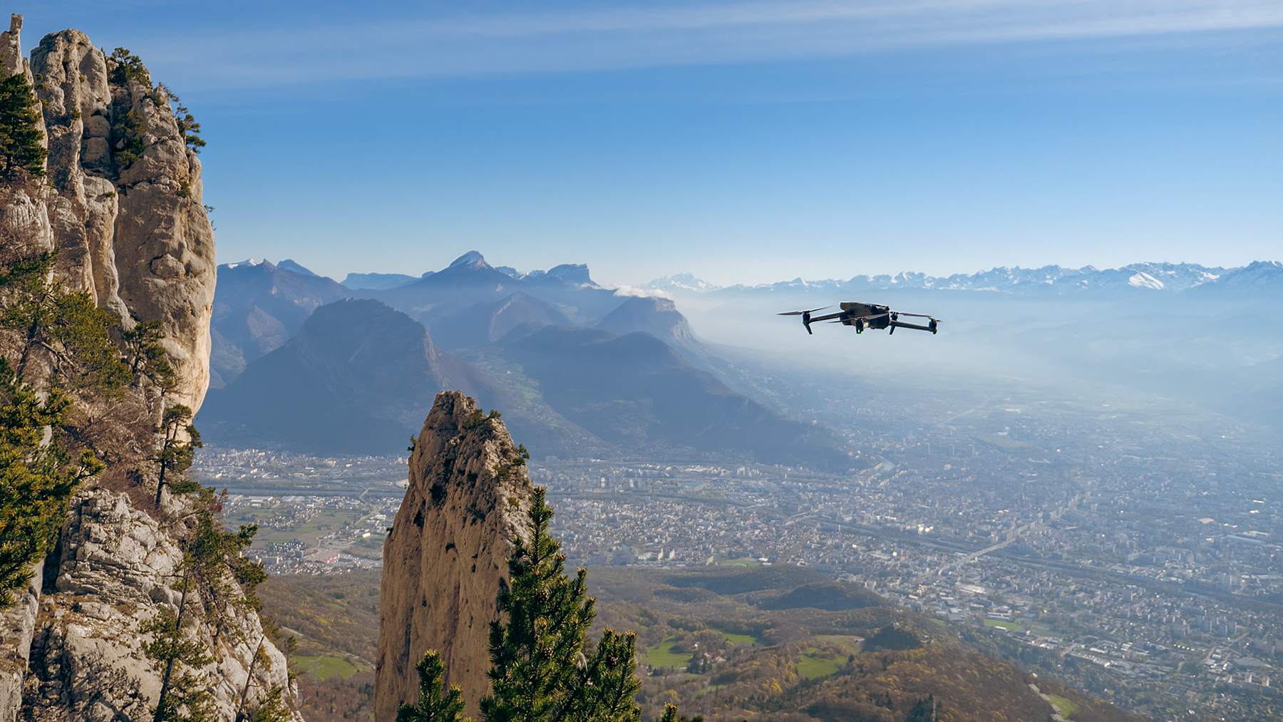 Un drone noir volant au-dessus d'une vallée avec des montagnes rocheuses et une ville en arrière-plan.