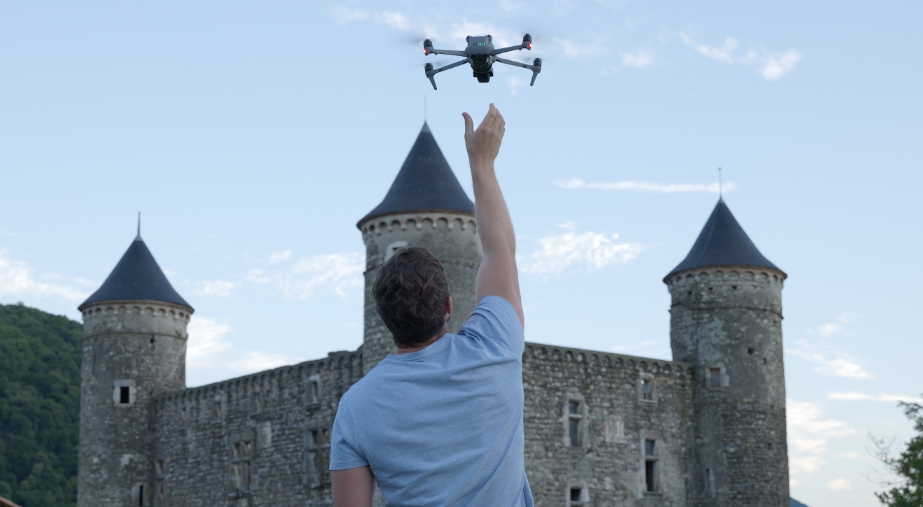 Un homme lève la main vers un drone volant devant un château en pierre aux tours coniques.