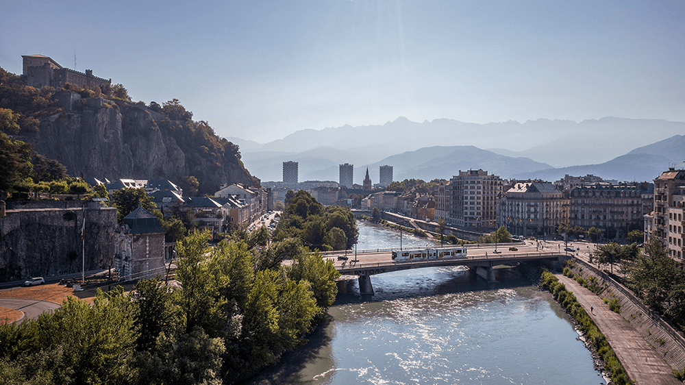 Vue panoramique de Grenoble avec la rivière, un pont, des immeubles et les montagnes en arrière-plan.